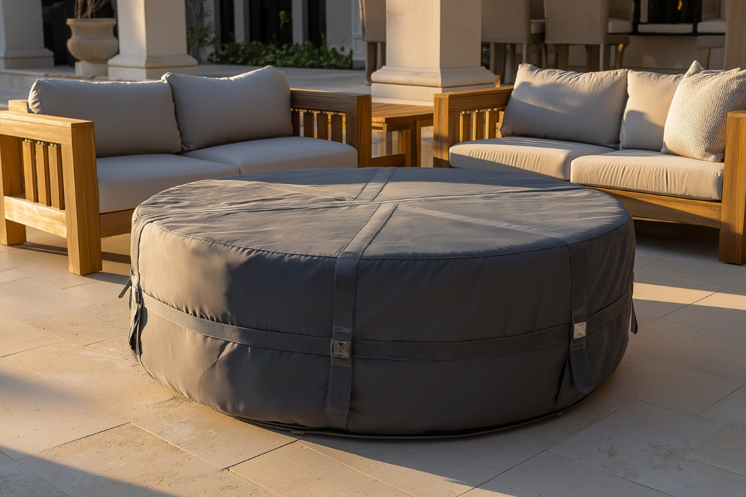 Gray round fire pit cover protecting a circular fire pit on a luxury patio, surrounded by teak wood outdoor sofas with light gray cushions in a sunlit backyard setting.