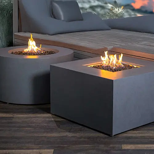 A dual pair of Beton concrete fire pits are on a patio deck ignited and displayed with lava rock.
