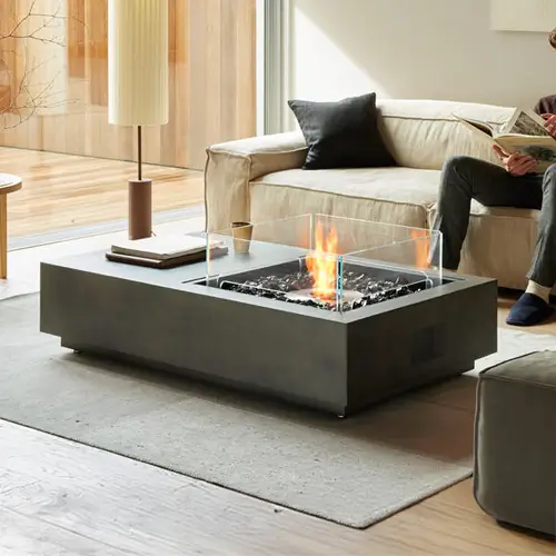 a man is reading a magazine in front of an ethanol concrete fire pit table set with several magazines and short books in his living room