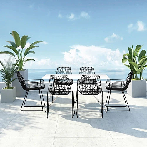 A modern outdoor dining set featuring a sleek white table with six black wireframe chairs, set against a stunning ocean view.