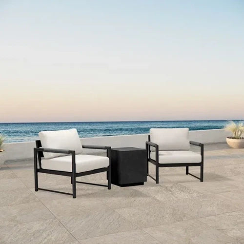 A modern outdoor lounge set with two black-framed club chairs featuring plush off-white cushions and a matching black side table, positioned on a beachfront patio.