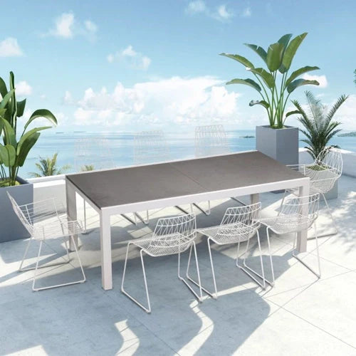 A modern outdoor dining set from the Harmonia Living Collection, featuring a large rectangular extension table with a dark grey surface and six sleek white wireframe chairs