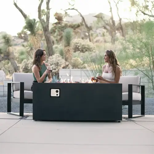 a pari of women enjoy the ambiance of the outdoor desert while dining and relaxing in front of a dark gray fire pit aptio set by the pool