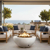 On an elevated terrace overlooking the ocean, a white concrete fire pit shaped like a smooth fire bowl burns with a bright ring of flames above dark lava rocks, centered between pale wood sofas with white cushions and navy pillows, while potted orange trees, grasses, and an outdoor grill line the back of the space, creating an outdoor fire pit lounge that feels like a relaxed backyard oasis with distant coastal views.