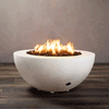A white half-sphere concrete fire pit sits on a striped wood-look floor in front of a plain taupe wall, the shallow fire bowl filled with chunky brown lava rocks while tall yellow flames from the gas fire pit rise and sway in the middle, clearly showing the rounded profile, narrow rim, small feet lifting it slightly from the ground, and a discreet control opening near the base.