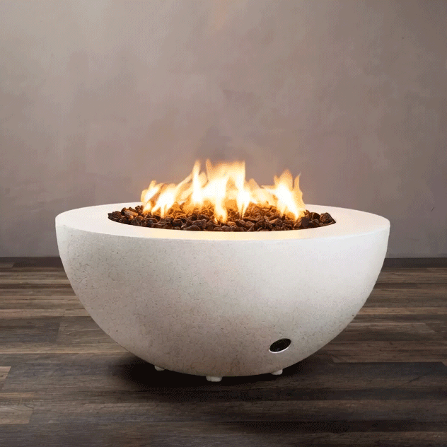 A white half-sphere concrete fire pit sits on a striped wood-look floor in front of a plain taupe wall, the shallow fire bowl filled with chunky brown lava rocks while tall yellow flames from the gas fire pit rise and sway in the middle, clearly showing the rounded profile, narrow rim, small feet lifting it slightly from the ground, and a discreet control opening near the base.