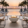 At a waterfront pool deck during sunset, a low white fire bowl acts as a round fire pit between modern lounge chairs and small metal side tables holding drinks, towels, and candles, its flames flickering over lava rocks while palm trees in large planters frame an infinity pool that appears to merge with the calm water beyond, illustrating an outdoor fire pit setting arranged for relaxed resort-style outdoor entertaining.