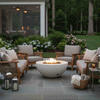 In a lush garden courtyard, wicker and wood lounge chairs with soft gray cushions encircle a white round fire pit on a slate patio, its compact flame glowing above lava rocks as lanterns, potted flowers, and tall trees surround the seating area, showing a patio fire pit layout that reads as a cozy backyard fire pit and natural family gathering fire pit spot just steps from the open doors of the house.