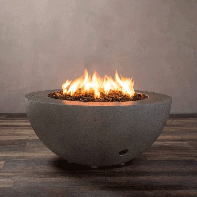 A low half-sphere fire bowl in warm brown textured concrete sits on a striped wood-look floor in front of a plain taupe wall, its wide rim framing a shallow basin filled with chunky lava rocks while bright yellow flames from the gas fire pit rise and bend at the center, clearly revealing the smooth curved profile, small feet lifting the concrete fire pit off the ground, and a discreet control port near the base.