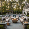 In a landscaped backyard with tall trees, stone planters, and a rectangular pool, two long sofas and matching armchairs form a U-shaped seating area around a brown concrete fire bowl, its round opening filled with glowing rocks and dancing flames, while lanterns, throw blankets, and a pergola-covered grill station complete the view, creating a cozy backyard fire pit scene that feels like a private backyard oasis for lingering chats and cool-night relaxation.