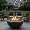 On a slate patio in front of a Tudor-style home, a low graphite fire bowl burns with tall yellow flames above lava rocks, encircled by cushioned wood chairs and a bench, with lanterns on the coffee table and a basket of blankets nearby, creating a patio fire pit vignette where the warm glow of this cozy backyard fire pit naturally invites guests to settle in, chat, and use it as a relaxed family gathering fire pit on cool evenings.