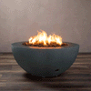 Indoors on a striped wood-look floor, a graphite gray concrete fire pit shaped like a deep fire bowl rests slightly lifted on small feet, its wide rim framing a recessed basin packed with chunky lava rocks while bright flames from the gas fire pit rise and sway in the center, clearly revealing the curved profile, textured surface, and discreet control port near the base against a simple taupe wall.