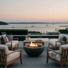 A graphite round fire pit in a smooth bowl design flickers at the center of a waterfront terrace, surrounded by teak lounge chairs with pale cushions, a low table set with candles and glasses, and neatly clipped hedges, while a calm pool, dock, and sailboats on still water reflect the soft pastel sky, turning this seating area into an outdoor fire pit retreat that doubles as an elegant backyard oasis by the bay.