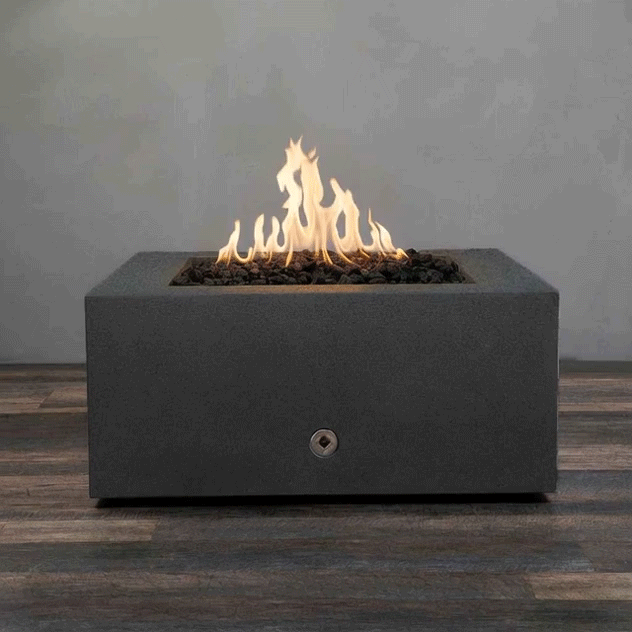 A charcoal-gray concrete fire pit with a low, boxy profile sits centered on weathered wood-look decking, its recessed square opening filled with rough black lava rocks while tall yellow flames rise and curl above them, a small round control set into the front panel, the square fire pit functioning as a compact gas fire pit that casts warm shifting light across the floorboards and up the plain gray wall behind it.