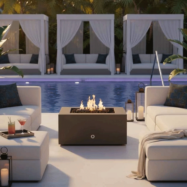 On a sleek poolside terrace at dusk, a graphite concrete fire pit glows at the center of white cushioned sectionals, its lively flames lifting from dark stones while low side tables hold drinks and lanterns and three curtained cabanas line the far edge of the blue water, the rectangular box serving as both a functional fire table and a luxury fire pit that anchors intimate outdoor entertaining in a lush palm-filled setting.