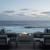 On a rooftop-style deck overlooking rolling ocean waves, a rectangular concrete fire pit glows at the center of a seating area with low gray loungers and slim metal side tables, the flames rising through dark stones while a glass railing frames the horizon, presenting a concrete fire pit used as a sleek fire table and modern fire pit that anchors relaxed outdoor entertaining in the soft blue evening light.