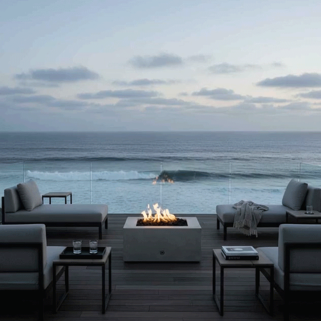 On a rooftop-style deck overlooking rolling ocean waves, a rectangular concrete fire pit glows at the center of a seating area with low gray loungers and slim metal side tables, the flames rising through dark stones while a glass railing frames the horizon, presenting a concrete fire pit used as a sleek fire table and modern fire pit that anchors relaxed outdoor entertaining in the soft blue evening light.