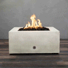A low white concrete fire pit with a clean square shape sits centered on dark wood-look decking, its recessed opening filled with rough black lava rocks as tall yellow flames rise and sway against a plain gray wall, the simple metal control on the front panel hinting at a tidy gas fire pit design that focuses attention on the glowing core of this compact square fire pit in an otherwise empty room.