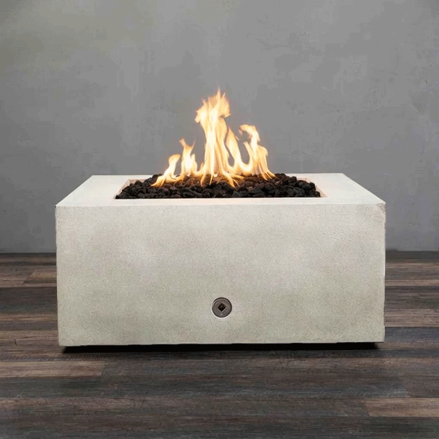 A low white concrete fire pit with a clean square shape sits centered on dark wood-look decking, its recessed opening filled with rough black lava rocks as tall yellow flames rise and sway against a plain gray wall, the simple metal control on the front panel hinting at a tidy gas fire pit design that focuses attention on the glowing core of this compact square fire pit in an otherwise empty room.