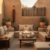 In a small stucco-walled courtyard with warm adobe tones, a square white concrete fire pit burns steadily between built-in bench seating layered with neutral cushions and patterned pillows, surrounded by terracotta pots, candles, cactus, and a hanging string of dried peppers, forming a snug patio fire pit that feels like a cozy backyard fire pit arranged for intimate outdoor entertaining in a Southwestern-inspired nook.