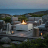 A white square concrete fire pit glows on a raised wooden deck perched above rocky coastal hills, its flames rising through dark stones as gray cushioned chairs and sofas circle around with chunky knit throws, lanterns, and a mug on a stump-style side table, depicting an outdoor fire pit that functions as a cozy backyard fire pit and inviting family gathering fire pit against a soft blue twilight sky.
