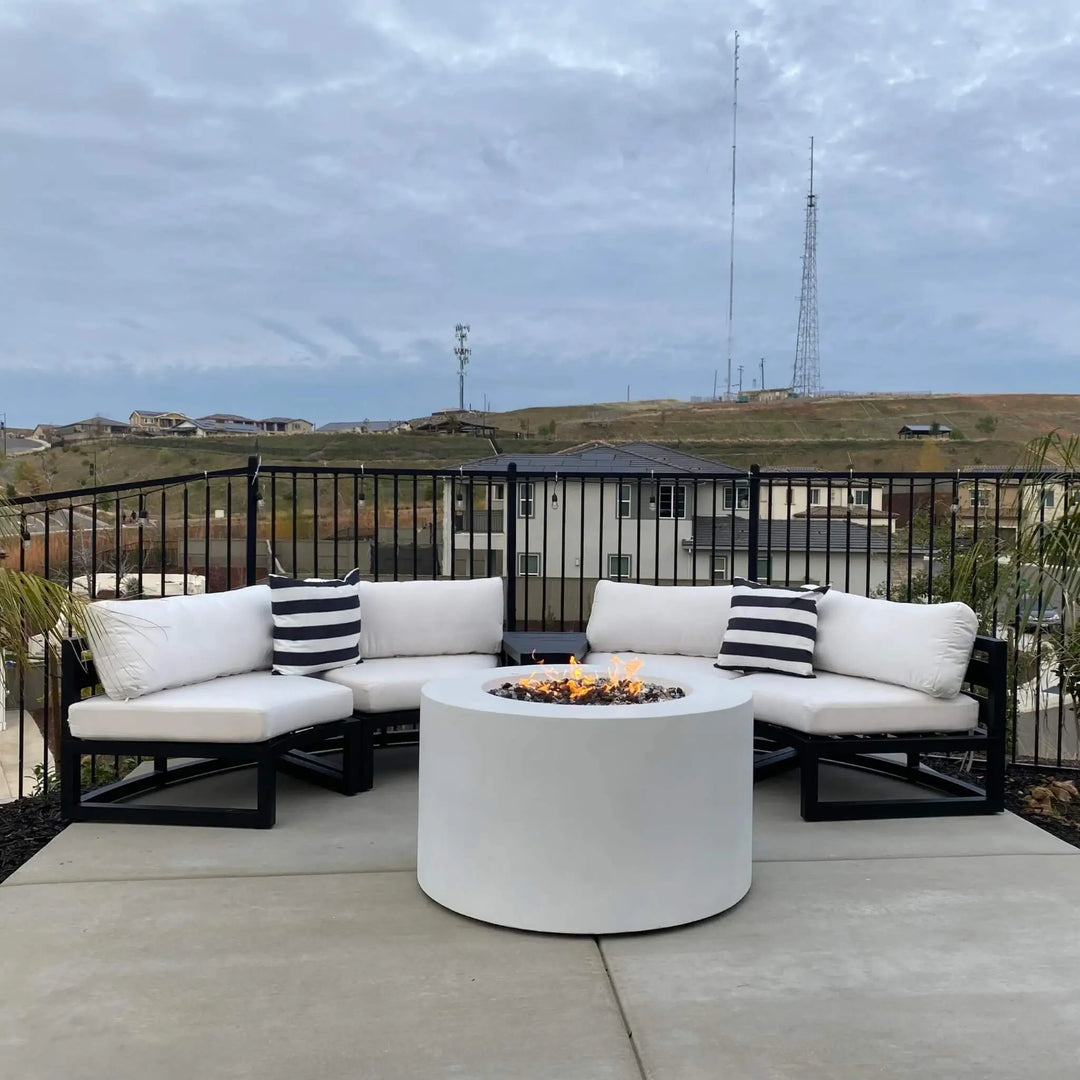 Modern white round GFRC concrete gas fire pit with hidden propane tank, lit and glowing with vibrant flames, centered on a spacious patio surrounded by cushioned white sectional seating and black striped pillows / perfect for cozy outdoor gatherings and backyard entertaining.