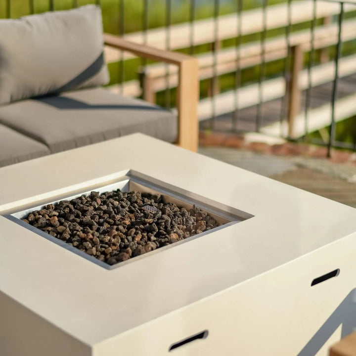 A close-up view of the 42" Square Beton Concrete GFRC Gas Fire Pit by Starfire Designs in a sleek white finish, shown without the flame to highlight its modern design and natural black lava rock media. The smooth GFRC surface and integrated propane tank storage create a clean, contemporary look, perfect for coastal decks, patios, or backyard lounging areas. This image showcases the fire pit's premium craftsmanship and durable materials, ideal for homeowners seeking a minimalist outdoor heating solution.