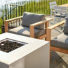 Close-up of a 42" square white GFRC concrete gas fire pit by Starfire Designs, featuring a recessed burner pan filled with lava rock, surrounded by modern teak and cushion lounge chairs on a sunlit patio with scenic lake views in the background.