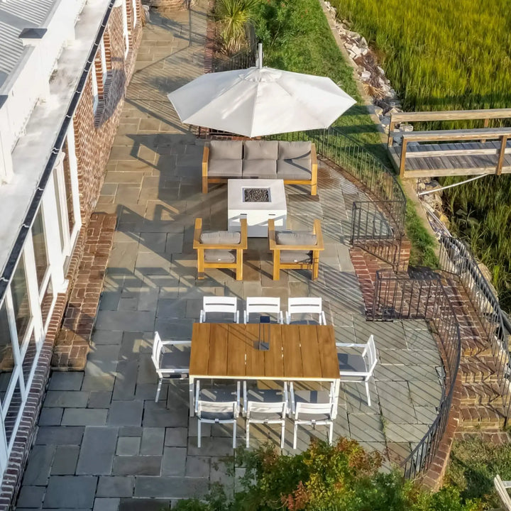 Aerial view of a coastal patio showcasing the 42" Square Beton Concrete GFRC Gas Fire Pit in White by Starfire Designs, elegantly paired with modern teak lounge chairs under a large white umbrella. The clean lines of the fire pit complement the waterfront setting, surrounded by lush greenery, a dock, and a spacious outdoor dining area with white and natural wood accents / perfect for entertaining or unwinding outdoors in style.