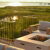 A crisp white 42-inch square Beton concrete gas fire pit by Starfire Designs, featuring a hidden propane tank and modern lava rock burner, radiating a cozy flame as it anchors a stylish outdoor seating area. The serene waterfront view and golden hour glow highlight the luxurious yet inviting atmosphere / perfect for evening relaxation or entertaining on the patio.