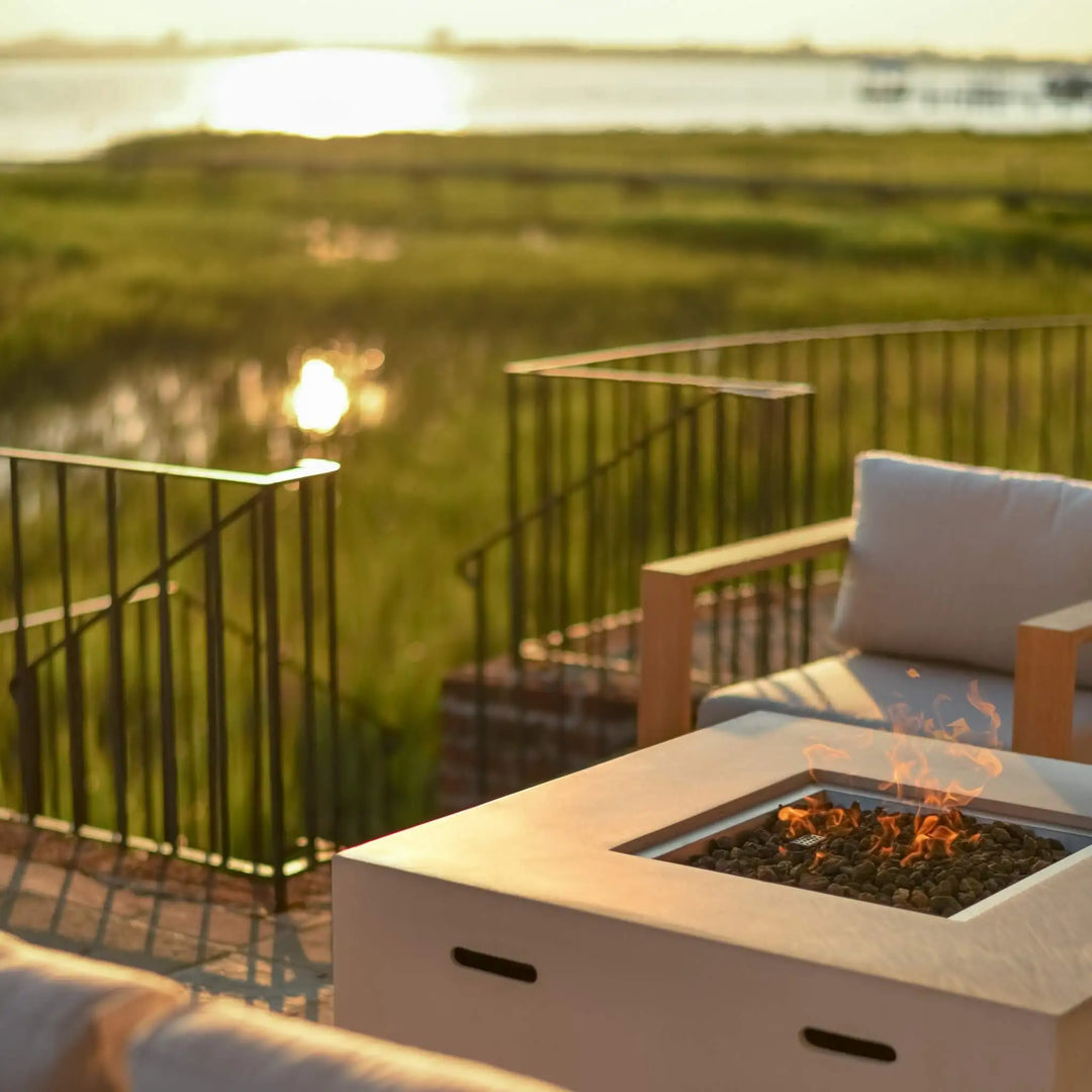 A crisp white 42-inch square Beton concrete gas fire pit by Starfire Designs, featuring a hidden propane tank and modern lava rock burner, radiating a cozy flame as it anchors a stylish outdoor seating area. The serene waterfront view and golden hour glow highlight the luxurious yet inviting atmosphere / perfect for evening relaxation or entertaining on the patio.