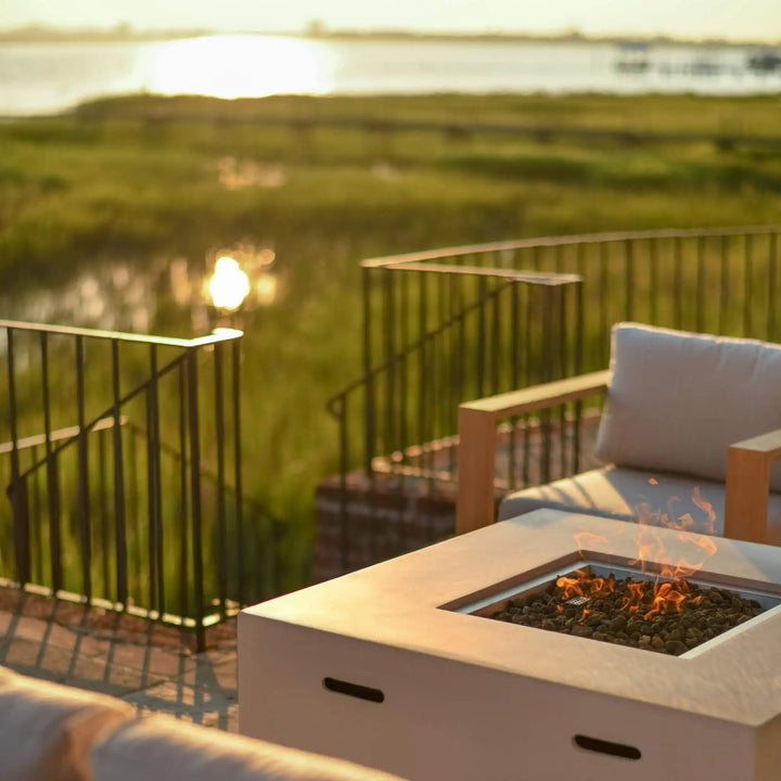 A crisp white 42-inch square Beton concrete gas fire pit by Starfire Designs, featuring a hidden propane tank and modern lava rock burner, radiating a cozy flame as it anchors a stylish outdoor seating area. The serene waterfront view and golden hour glow highlight the luxurious yet inviting atmosphere / perfect for evening relaxation or entertaining on the patio.