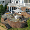 Aerial view of a stunning patio setup featuring the 42-inch Square Beton Concrete GFRC Gas Fire Pit in White by Starfire Designs. Positioned at the heart of a curved brick terrace overlooking the water, this sleek fire pit with hidden propane tank is surrounded by stylish teak and charcoal lounge seating under a large cantilever umbrella. Ideal for entertaining or relaxing outdoors, this luxury fire feature adds modern elegance to a serene waterfront backyard.