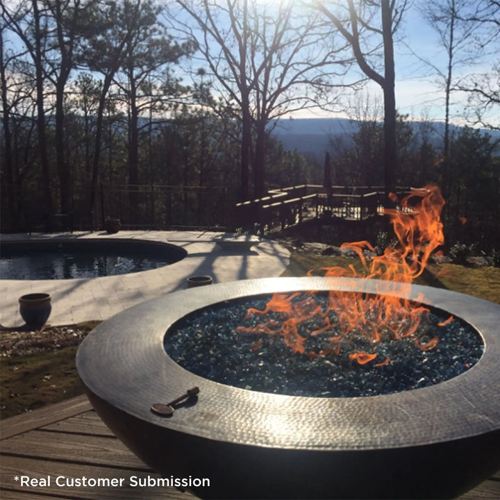 Customer photo of a round copper fire pit with blue fire glass, set on a wooden deck overlooking a scenic forest view and a swimming pool, with flames dancing under a clear blue sky