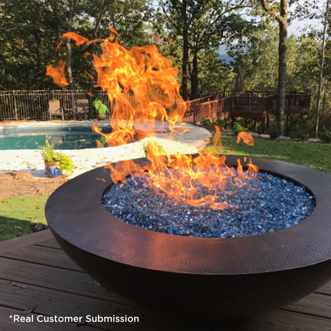 Customer photo of a round copper fire pit filled with blue fire glass, positioned on a wooden deck near a pool, with bright flames rising against a backdrop of lush greenery and trees