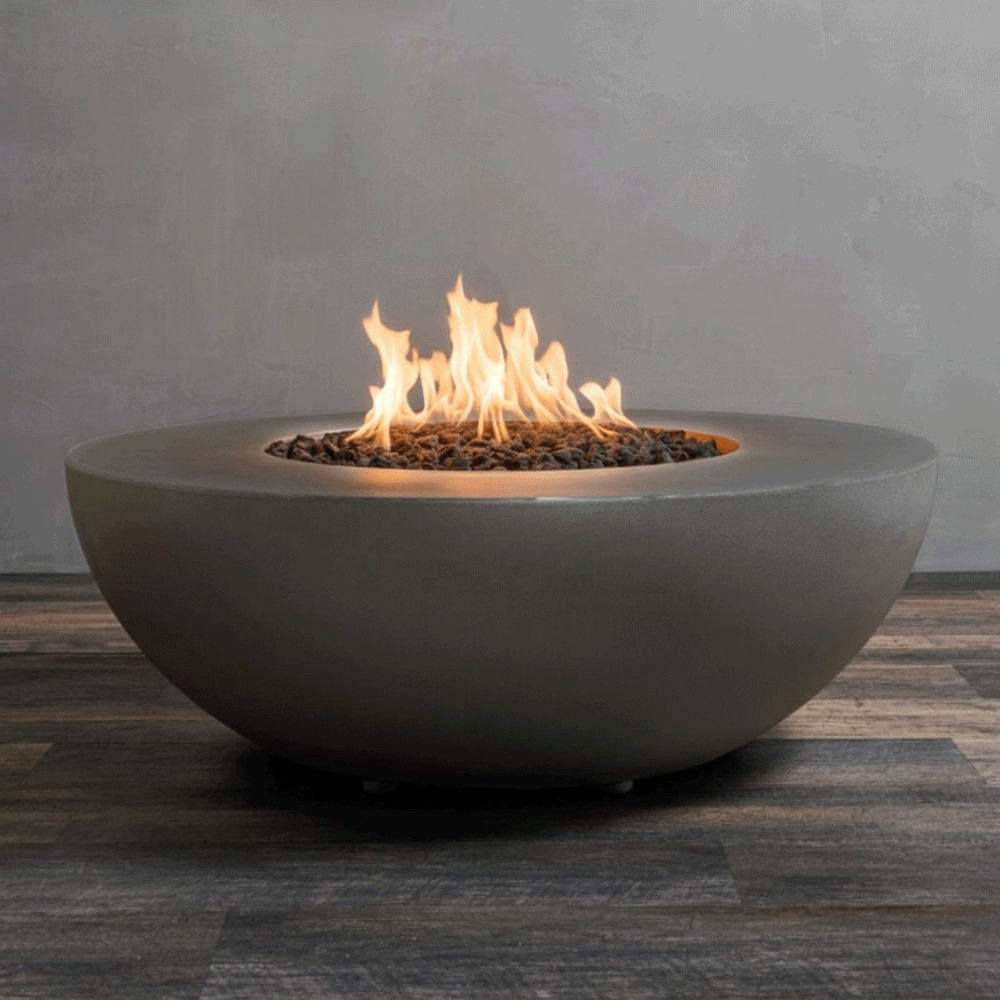 A studio close-up features a round fire pit in a rich brown finish, resting on a dark wood floor. Bright yellow flames burn centrally from a bed of black lava rocks, highlighting the curved profile of the concrete fire pit. This minimalist view emphasizes the earthy tone and modern sculptural design of the fire bowl, showcasing the clean combustion of the natural gas fire pit unit.