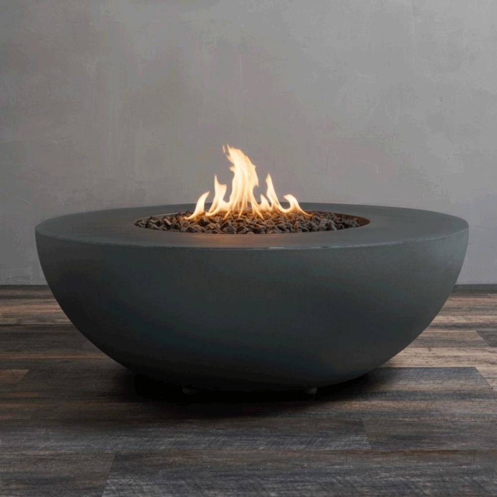 A studio presentation highlights a fire bowl in a matte graphite finish, resting on a dark wood floor against a grey wall. Bright orange flames rise centrally from a bed of black lava rocks, showcasing the smooth texture and modern profile of the concrete fire pit. This view emphasizes the clean combustion and minimalist design of the natural gas fire pit unit.