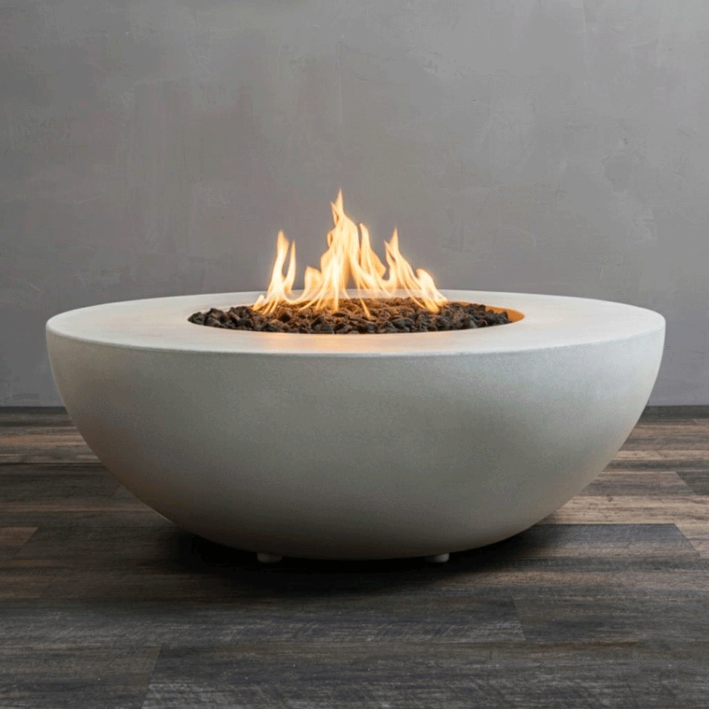 A studio close-up features a round fire pit in a crisp white finish, resting on a dark wood floor. Bright yellow flames burn steadily from a bed of black lava rocks, highlighting the smooth curve of the concrete fire pit. This minimalist presentation emphasizes the clean lines and modern aesthetic of the fire bowl, demonstrating the efficient operation of a natural gas fire pit connection.