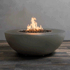 A low, round fire pit made from smooth taupe concrete sits centered on a plank-style floor, its wide bowl filled with uneven dark lava rocks as a tall golden flame quietly rises and shifts in the breeze, highlighting a concrete fire pit designed as a clean, modern gas fire pit that casts a soft amber glow across the rounded rim, the textured wood surface below, and the plain gray wall behind it in a simple, unfurnished setting.