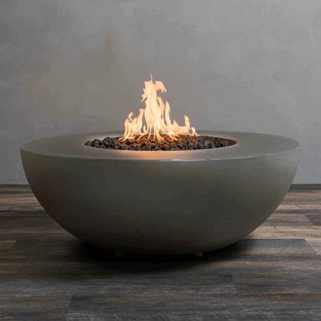 A low, round fire pit made from smooth taupe concrete sits centered on a plank-style floor, its wide bowl filled with uneven dark lava rocks as a tall golden flame quietly rises and shifts in the breeze, highlighting a concrete fire pit designed as a clean, modern gas fire pit that casts a soft amber glow across the rounded rim, the textured wood surface below, and the plain gray wall behind it in a simple, unfurnished setting.