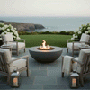 A circular concrete fire pit burns with a steady golden flame on a gray stone patio above a manicured green lawn and distant ocean cliffs, surrounded by four cushioned wooden lounge chairs, blooming white hydrangea bushes, side tables, and metal lanterns, creating an outdoor fire pit that anchors the seating area and defines a tranquil backyard oasis ideal for relaxed conversation and casual outdoor entertaining as daylight fades.
