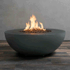 A deep graphite fire bowl made of smooth concrete sits on a striped wood-look floor in front of a plain gray wall, the wide circular rim framing a recessed basin filled with chunky lava rocks while tall yellow flames from the gas fire pit rise and flicker in the center, clearly showing the round profile, low stance, and even glow of this compact concrete fire pit as it burns in place.