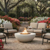 In a flowering garden bordered by azaleas and moss-draped trees, four metal-framed chairs and a small sofa form a loose ring around a pale concrete round fire pit set on gravel, its compact flame glowing over a bed of stones while a wrought-iron gate and brick path extend into the background, presenting a backyard fire pit nook that feels like a cozy backyard fire pit and easy family gathering fire pit for quiet evenings outside.