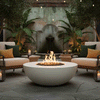 A smooth white fire bowl burns with a lively circle of flames at the center of a stone courtyard, surrounded by cushioned wicker chairs, tall potted plants, and hanging string lights, while a weathered stucco wall with an arched fountain and broad tropical leaves fills the backdrop, turning the outdoor fire pit area into an intimate backyard oasis well suited to low-key outdoor entertaining with candles and drinks scattered around.