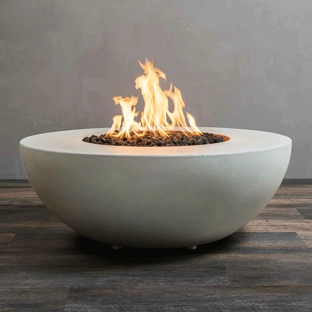 A smooth white fire bowl shaped like a wide, shallow half sphere sits on a striped wood-look floor against a plain gray wall, its recessed center filled with dark lava rocks while tall yellow flames from the gas fire pit rise and fold back on themselves, clearly outlining the round concrete fire pit’s low profile, broad rim, and soft matte texture as it appears to hover just above the ground.