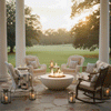 On a broad stone veranda framed by tall white columns and open lawn at sunset, wicker armchairs and rocking chairs with striped cushions form a loose circle around a low white round fire pit, its even ring of flame shining over small stones, with lanterns, quilts, and drinks resting on nearby tables, creating a cozy backyard fire pit arrangement that reads as a relaxed family gathering fire pit on a summer evening.