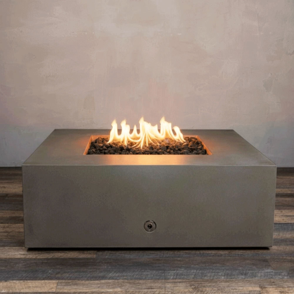 A studio close-up features a square fire pit in a rich brown finish, resting on a dark wood floor. Bright yellow flames burn continuously from a bed of black lava rocks, highlighting the clean lines and earthy tone of the concrete fire pit. The visible chrome key valve on the side indicates the manual control of this natural gas fire pit unit, demonstrating its simple and elegant functionality.