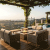 A brown luxury fire pit sits on a hillside wooden deck under a vine-covered pergola. Framed by comfortable wicker armchairs, the unit overlooks a city skyline and infinity pool at sunset. The steady flame of the patio fire pit creates a warm focal point for outdoor entertaining, with the gas fire pit providing ambiance in a scenic, elevated setting.