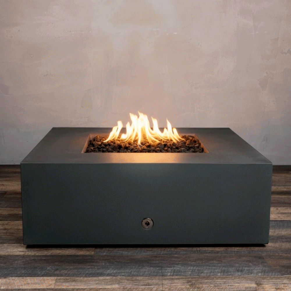 A studio presentation displays a square fire pit in a matte graphite finish, resting on a dark wood floor. Bright orange flames flicker continuously from a bed of black lava rocks, contrasting with the smooth texture of the concrete fire pit. The visible chrome key valve suggests a manual ignition system, demonstrating the clean design of this natural gas fire pit unit.