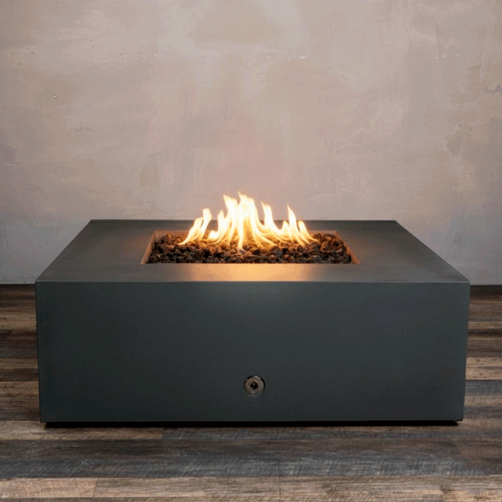 A studio presentation displays a square fire pit in a matte graphite finish, resting on a dark wood floor. Bright orange flames flicker continuously from a bed of black lava rocks, contrasting with the smooth texture of the concrete fire pit. The visible chrome key valve suggests a manual ignition system, demonstrating the clean design of this natural gas fire pit unit.